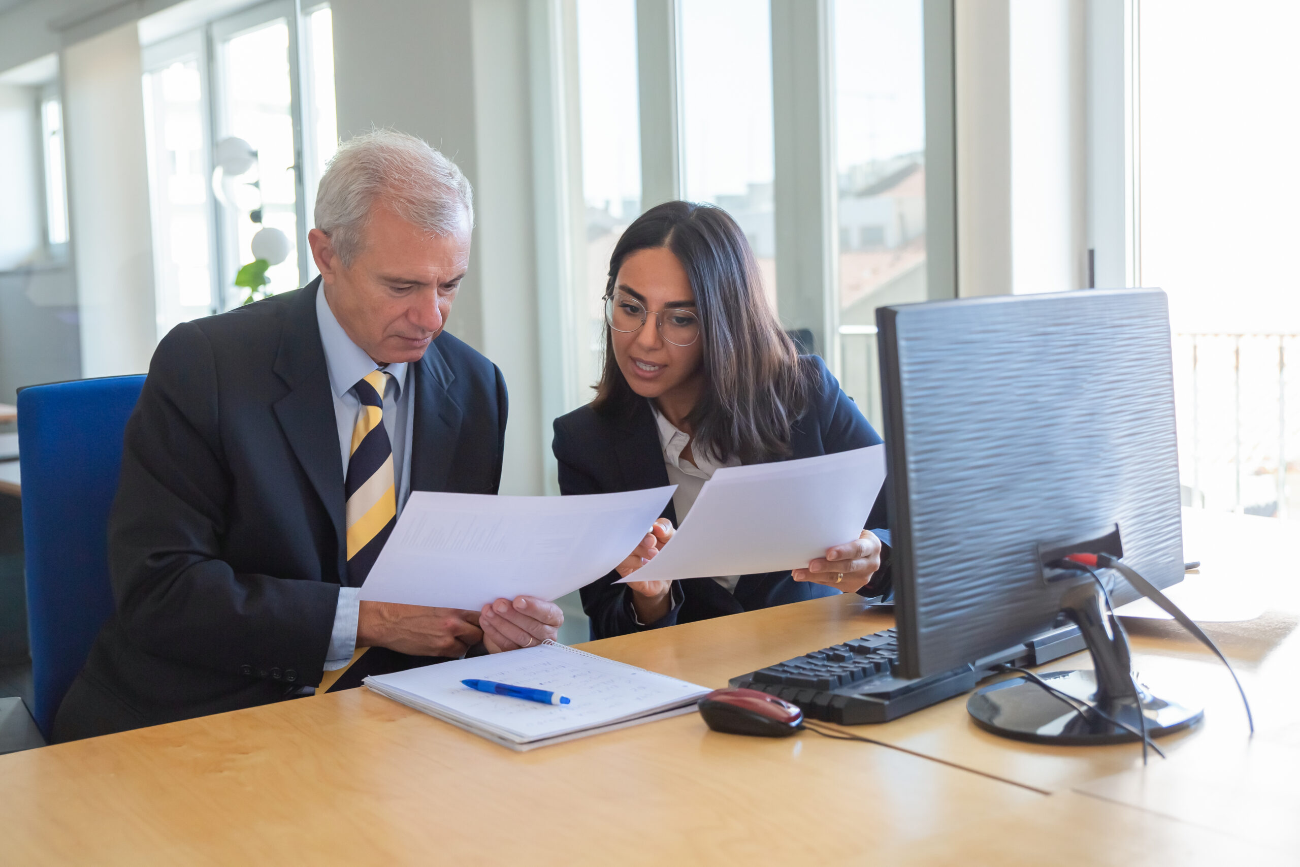 Profissional explicando detalhes de documentos para um cliente em um ambiente de trabalho. Ambos estão sérios e concentrados, simbolizando uma consultoria financeira ou legal, com foco em cooperação e trabalho em equipe.